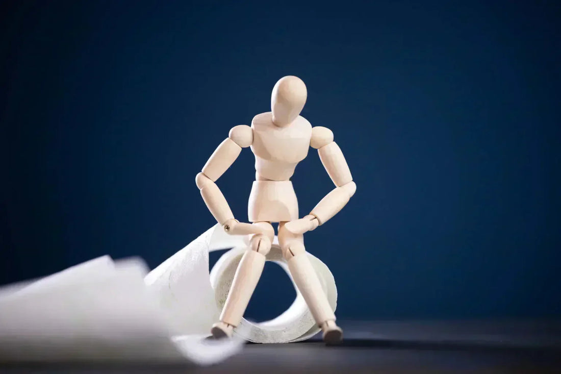 Ce petit bonhomme en bois se questionne sur les causes de la constipation, assis sur un rouleau de papier toilette