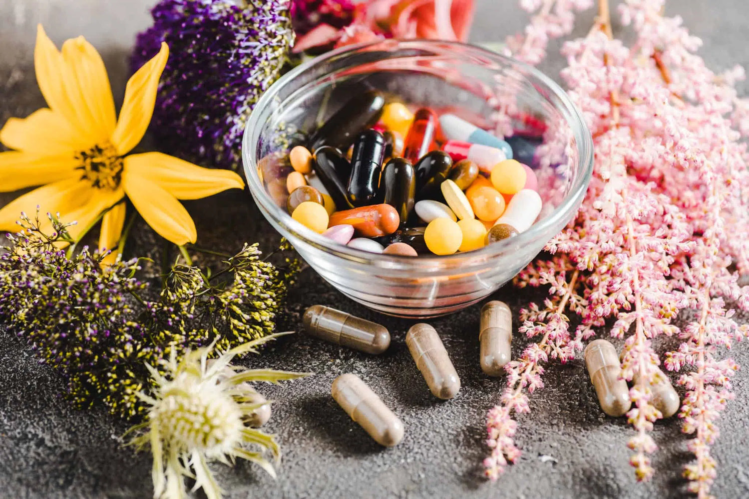 Une séléction des meilleurs actifs de santé naturels pour l'énergie, comme le montre ce bol transparent rempli de gélules et capsules, entouré de fleurs et de plantes médicinales