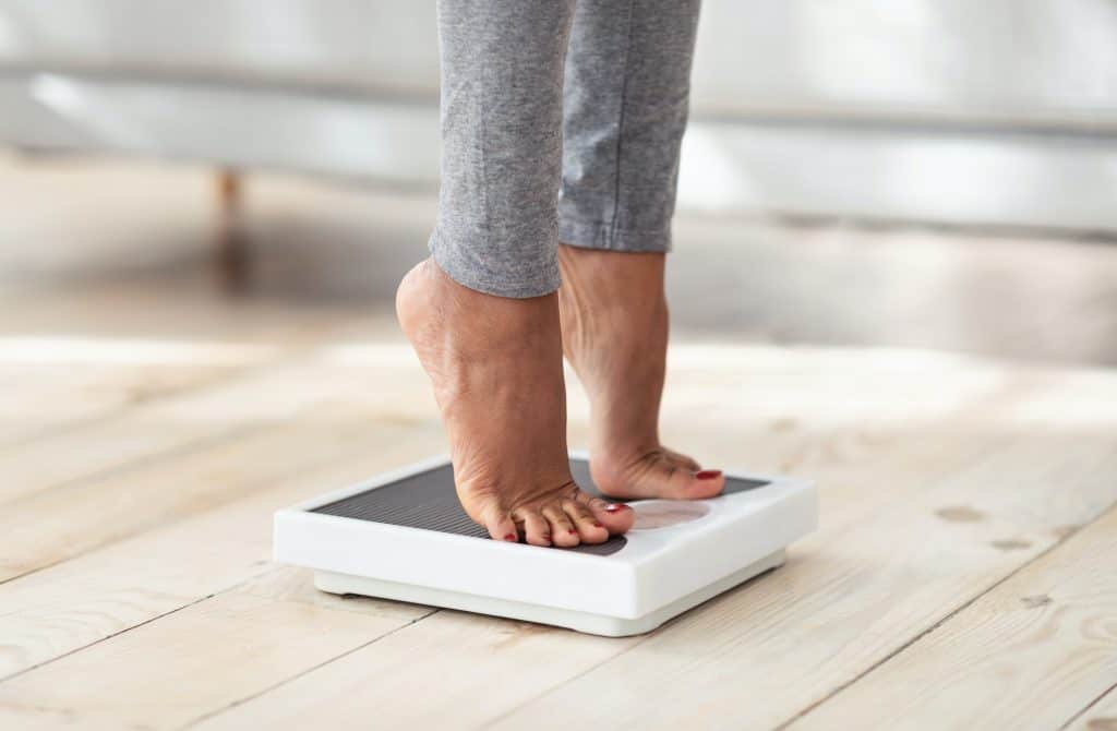 Femme vérifiant si les effets indésirables de la mélatonine sur l’augmentation du poids sont vérifiées en se pesant sur une balance du bout des pieds